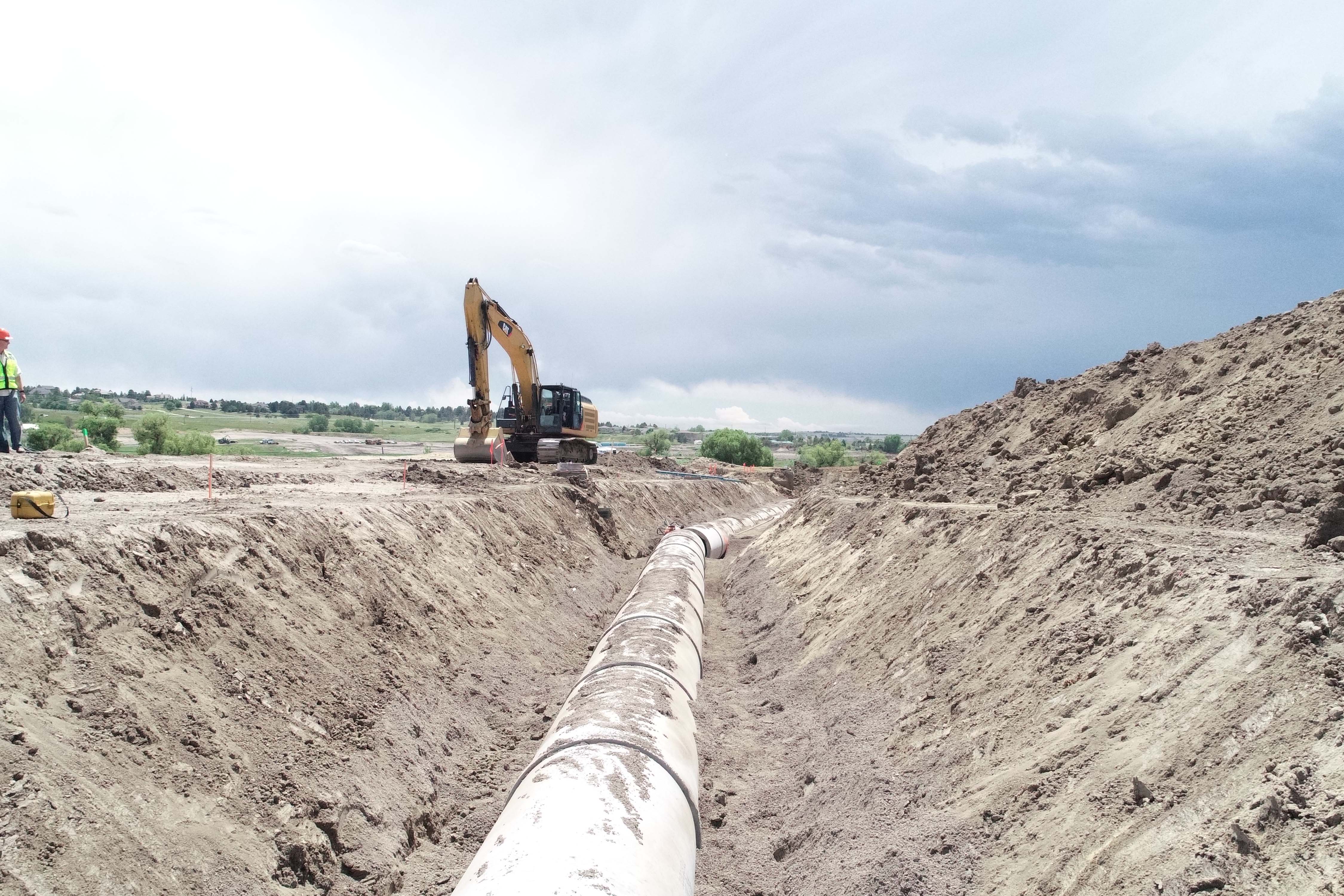 HEI Excavator in distance with construction worker on side, pipe in ground